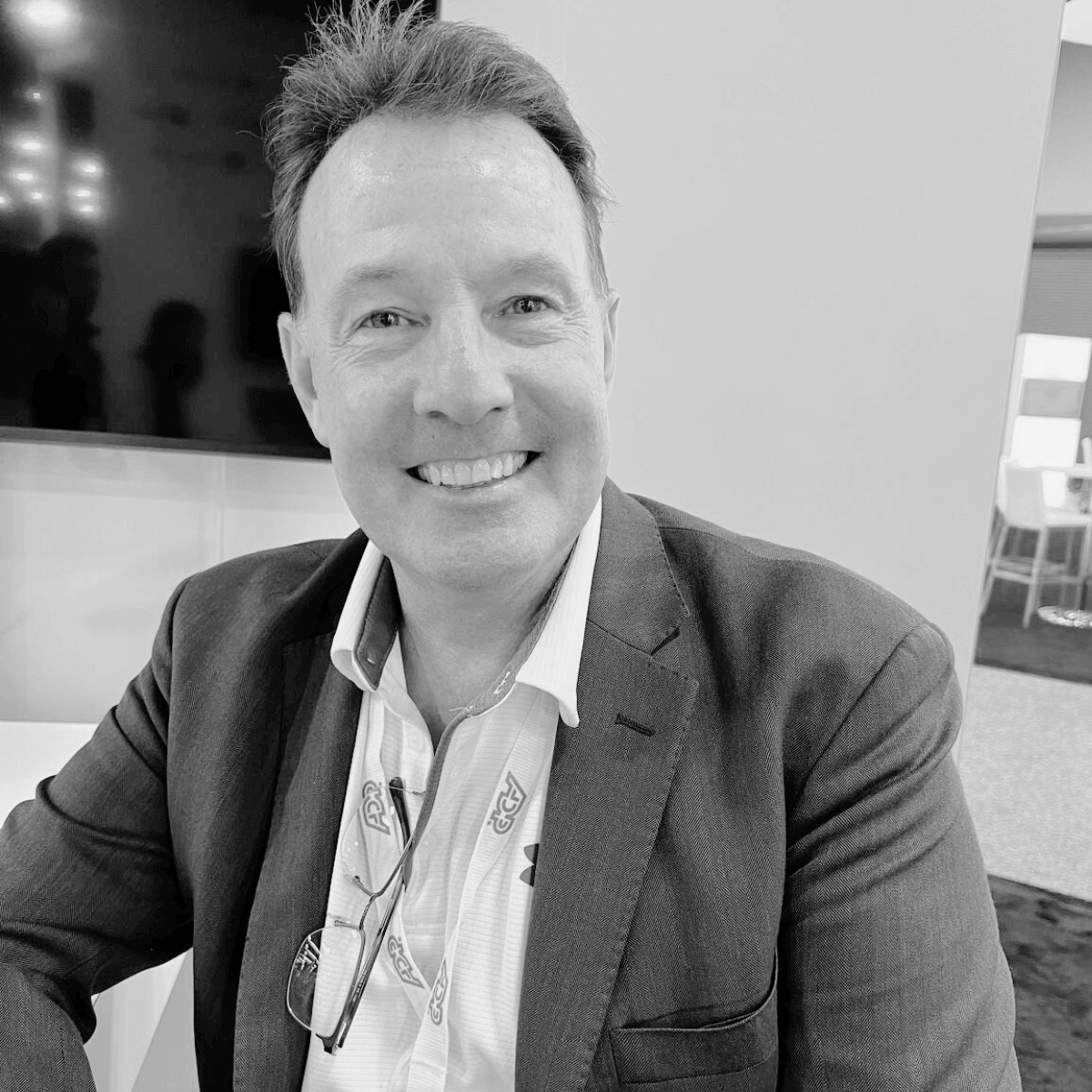 Smiling man in a suit and white shirt, seated indoors at an event. Background shows a large screen and modern furnishings. Black and white image.