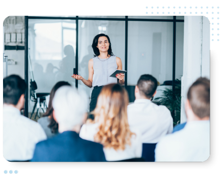 A woman confidently presents to an engaged audience in a modern office. She holds a tablet, with large windows and a glass partition behind her.